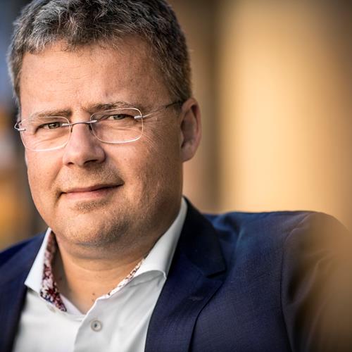 Man i kostym ler tittar mot kameran och kisar lite under sina glasögon