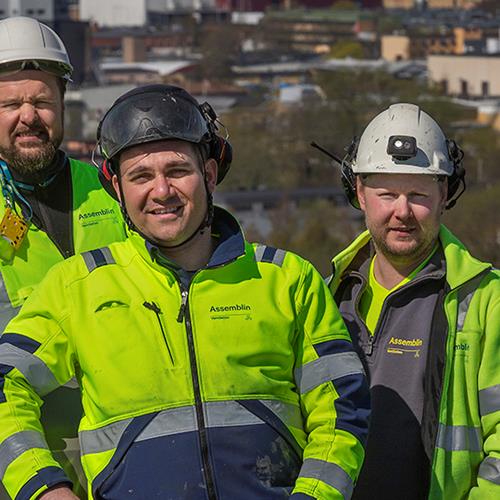 Tre män stående på hög höjd ler mot kameran i sina arbetskläder och hjälmar på huvudet